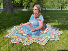 Load image into Gallery viewer, Woman sitting cross-legged on a pastel-striped crochet Superstar blanket spread on grass, with its star points fanning out around her.