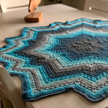 Load image into Gallery viewer, Low-angle view of a turquoise, aqua, navy, and gray crochet Supernova blanket spread on a light table, showing granny clusters and star points.

